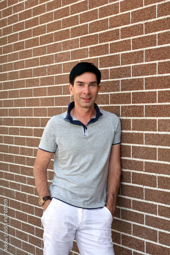 Счастливый улыбающийся уверенный в себе молодой человек в рубашку и белые летние брюки позирует возле кирпичной стены
