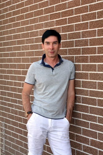 Счастливый улыбающийся уверенный в себе молодой человек в рубашку и белые летние брюки позирует возле кирпичной стены
