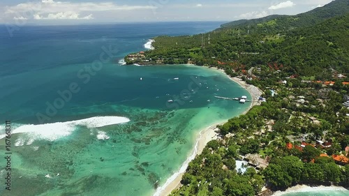 Aerial view of Senggigi beach with turquoise water in Lombok island covered by green trees, some hotels on the shore. Shot with drone on sunny day with blue sky in Indonesia, fly forward photo