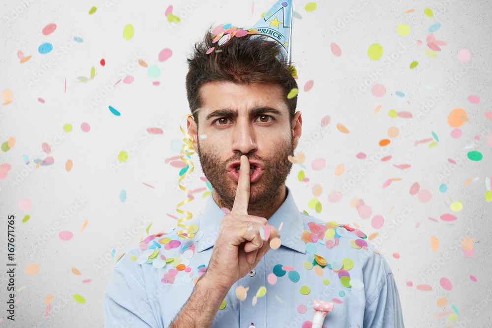 Φωτογραφία Attractive bearded young Caucasian male in blue shirt holding index finger at his lips, pronouncing Shh as he doesn't want to ruin surprise party on his girlfriend's birthday