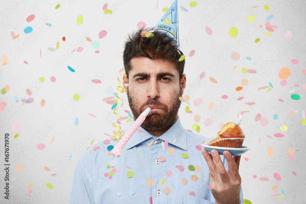 Φωτογραφία Displeased unhappy bearded Caucasian man with cone hat on head and party horn in mouth looking at camera with bored dissatisfied expression as his birthday party sucks