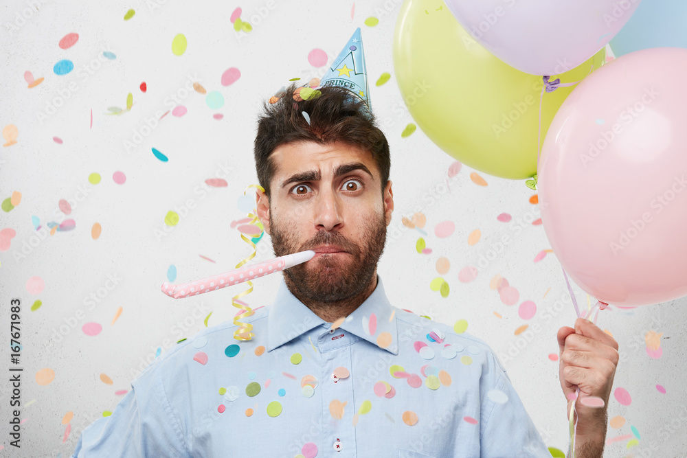 Φωτογραφία Indoor shot of astonished puzzled guy wearing blue shirt and cone hat on his head looking surprised, holding helium balloons and blowing on party horn