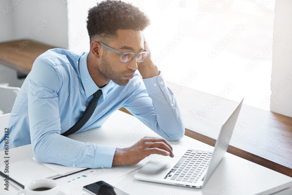 Candid shot of stylish young Afro American unshaven manager wearing formal shirt and glasses having bored look, wasting his time using laptop pc, browsing internet, doing nothing instead of working Candid shot of stylish young Afro American unshaven manager wearing formal shirt and glasses having bored look, wasting his time using laptop pc, browsing internet, doing nothing instead of working