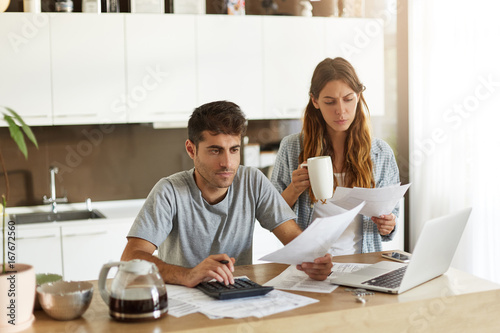 Picture of serious young American couple spending weekend morning in kitchen, doing paperwork, looking through mail and calculating family expenses, paying bills online using laptop computer © wayhome.studio 