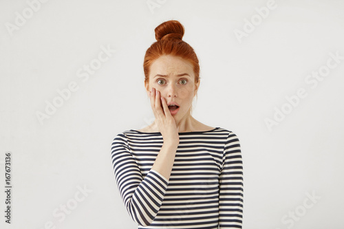 People, suddenness and surprise concept. Surprised female with red hair tied in bun, wearing striped sweater, keeping her hand on cheek, opening mouth. Redhead young girl with puzzled expression