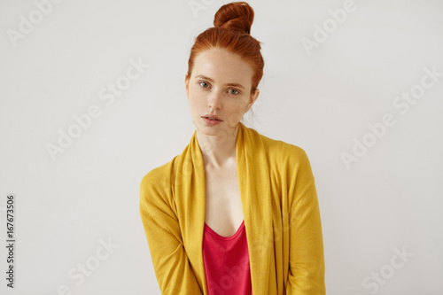 Serious assured good-looking girl, having red hair bun, freckled skin, thin lips and green eyes, wearing red shirt and yellow cape posing in studio against white background. Confidence and assuarance photo