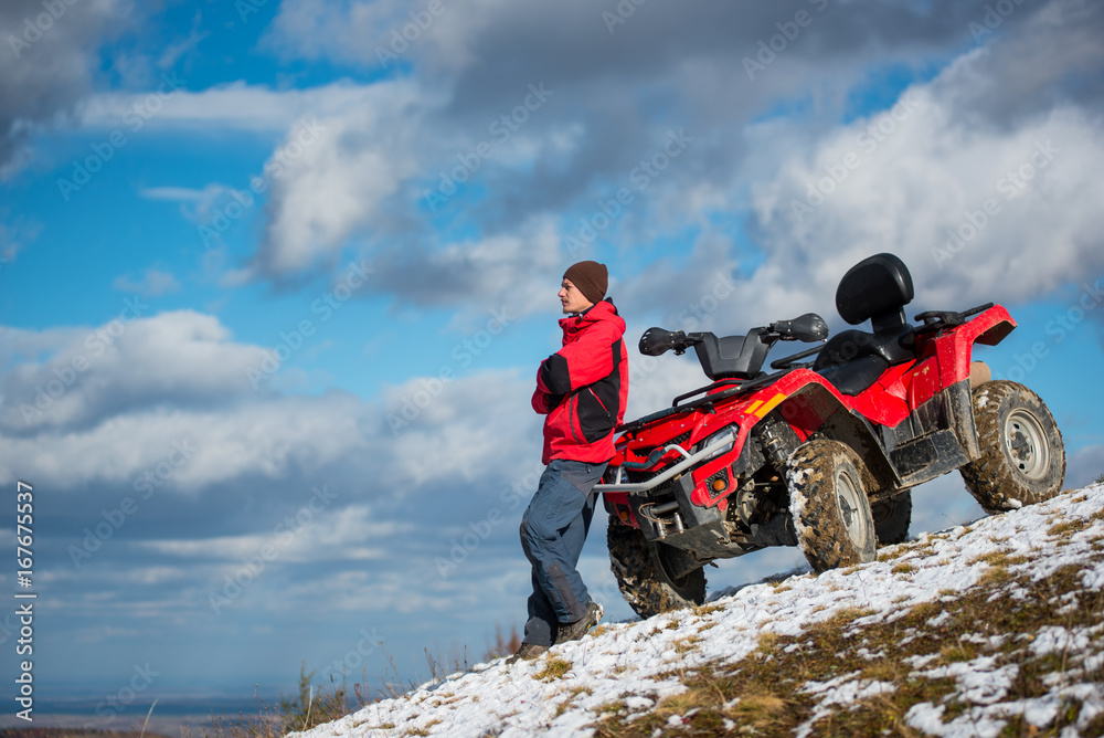 Φωτογραφία Guy in a red winter clothes standing near red ATV quad bike, looking into the distance on snowy mountain slope