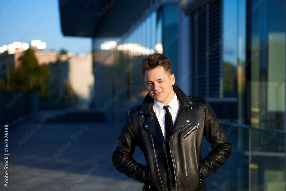 Lifestyle urban portrait of handsome young male employee in trendy leather jacket standing outside modern building with glass door, waiting for collegues for lunch, looking and smiling at camera
