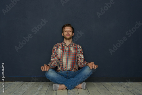 Casually-dressed bearded young Caucasian employee sitting on wooden floor at home in half-lotus posture, keeping eyes closed, meditating, relaxing body and mind after hard working day in office