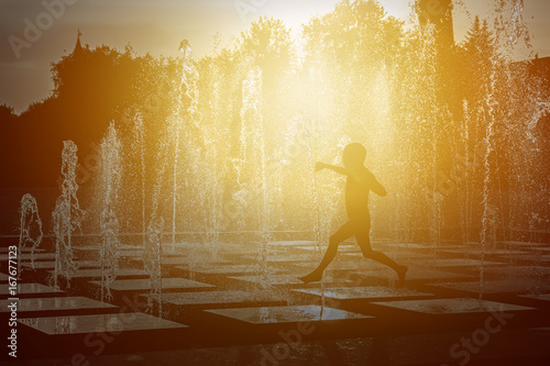 Fototapeta Naklejka Na Ścianę i Meble -  Silhouette of a child playing with a spray of a fountain on a hot summer day.