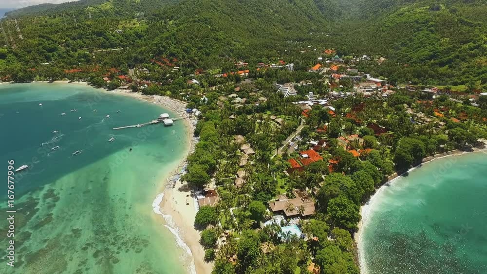 Aerial view of Senggigi beach with turquoise water in Lombok island covered by green trees, houses and hotels on the shore. Shot with drone on sunny day in Indonesia, fly forward