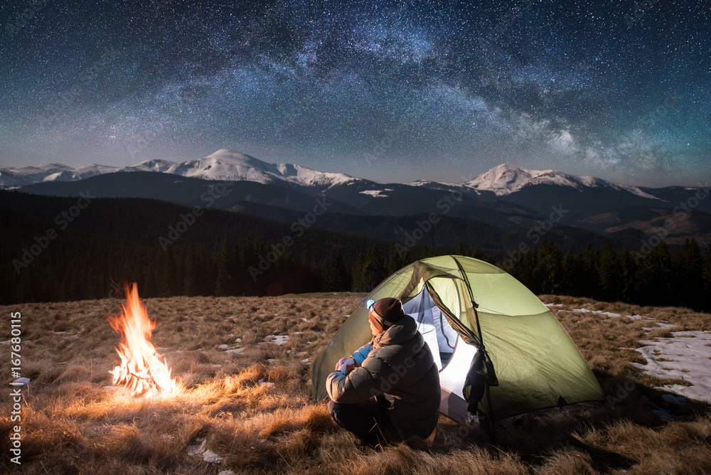 Fototapeta premium Male tourist have a rest in his camping in the mountains at night. Man with a headlamp sitting near campfire and tent under beautiful night sky full of stars and milky way, and enjoying night scene