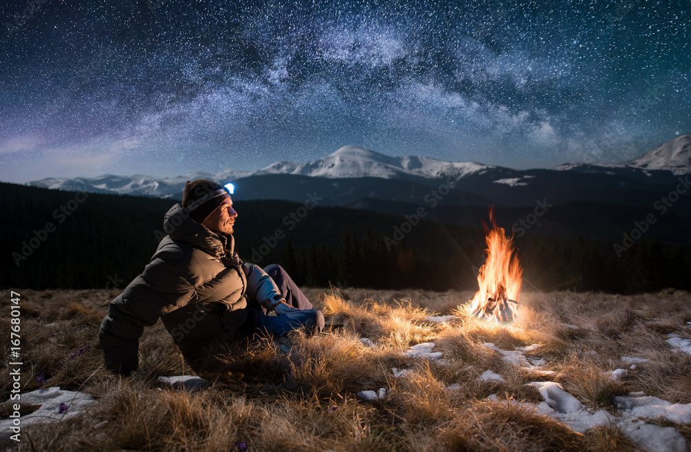 Male tourist have a rest in the mountains at night. Guy with a headlamp sitting near campfire under beautiful night sky full of stars and milky way, and enjoying night scene - obrazy, fototapety, plakaty 