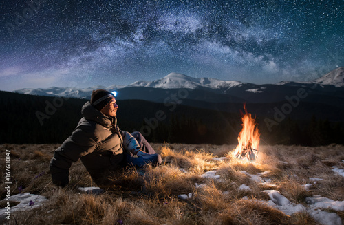 Male tourist have a rest in the mountains at night. Guy with a headlamp sitting near campfire under beautiful night sky full of stars and milky way, and enjoying night scene photo
