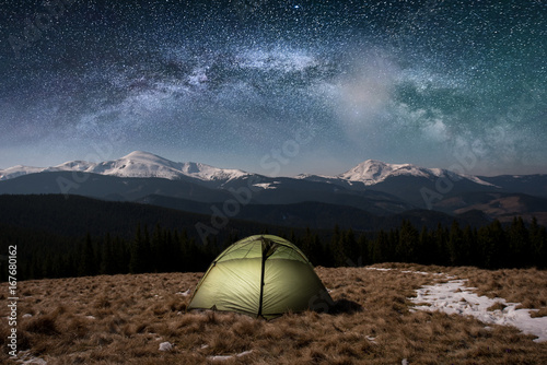 Fototapeta Naklejka Na Ścianę i Meble -  Night camping. Illuminated tourist tent under beautiful night sky full of stars and milky way. On the background snow-covered mountains and forests