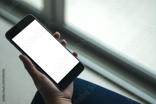 Mockup image of woman's hand holding black mobile phone with blank white screen on thigh with white tile floor and sliding door background