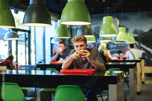 Fotografía Young beautiful student sitting in a fast food restaurant and eating an appetizing burger