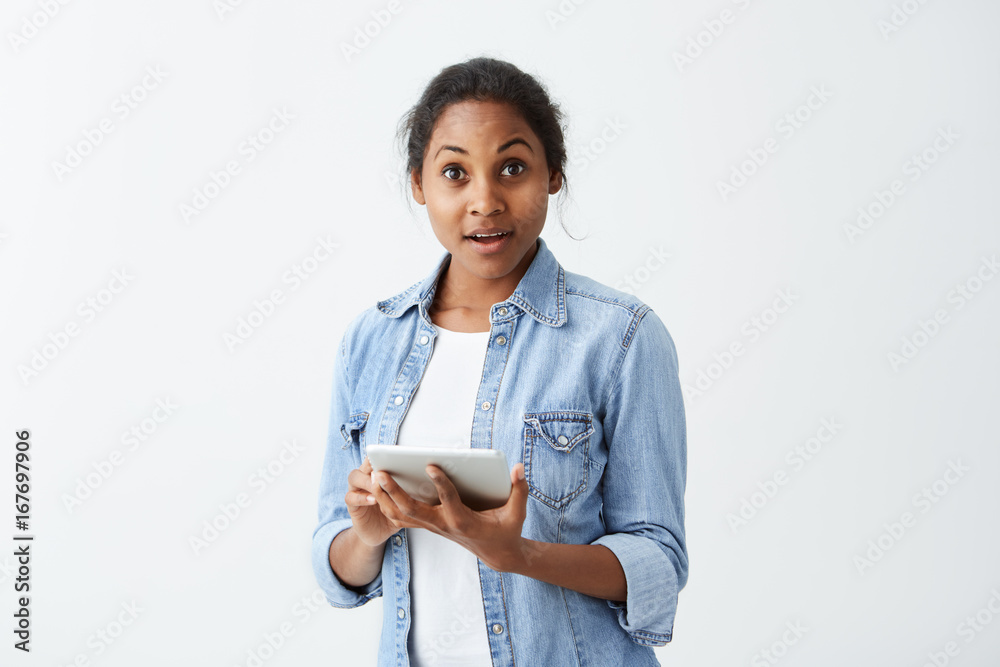 Forgetful Afro-American female with dark appealing in blue shirt holding tablet in her hands staring at camera with astonished amazed look, having completely forgotten about something serious. Forgetful Afro-American female with dark appealing in blue shirt holding tablet in her hands staring at camera with astonished amazed look, having completely forgotten about something serious.