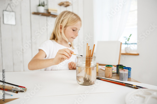 Сreative little blonde girl with freckles washing brush in jar of water during art lesson. Cute child concentrated on painting. Kids' art and activities.