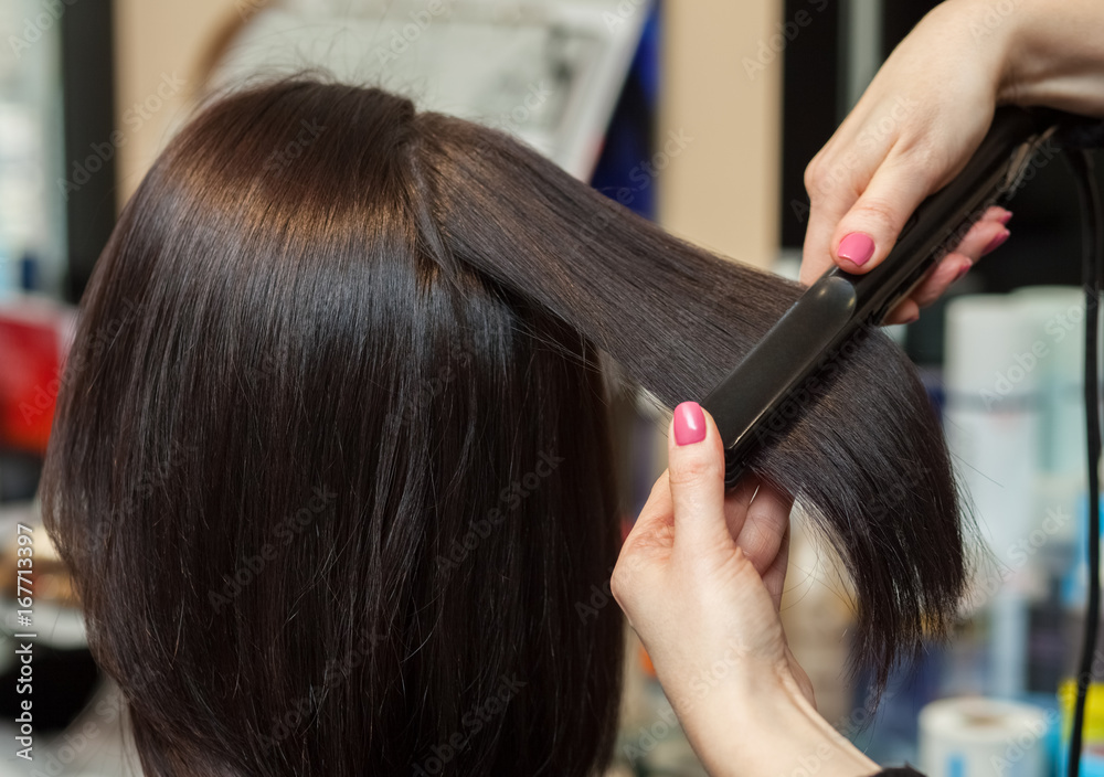 Fototapeta premium The hairdresser does aligns the hair with hair iron to a young girl, brunette in a beauty salon. Professional Hair Care