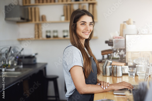 Young woman barista at her workplace looking happy smiling working on a laptop