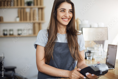Young female coffee shop employee using a credit card reader and a laptop to bill the customer