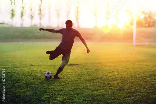 Soccer player is shooting a ball in stadium at sunset.