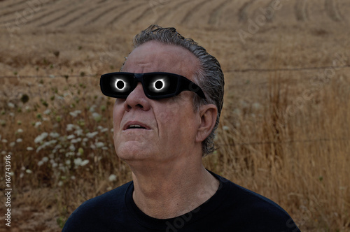 Man looking at full solar eclipse with eclipse reflecting in lenses/Man viewing full solar eclipse with solar glasses in country field/