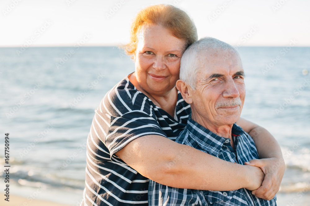 Happy family of two mature people hugging outdoors. Senior smiling couple standing on beach near sea on sunset.