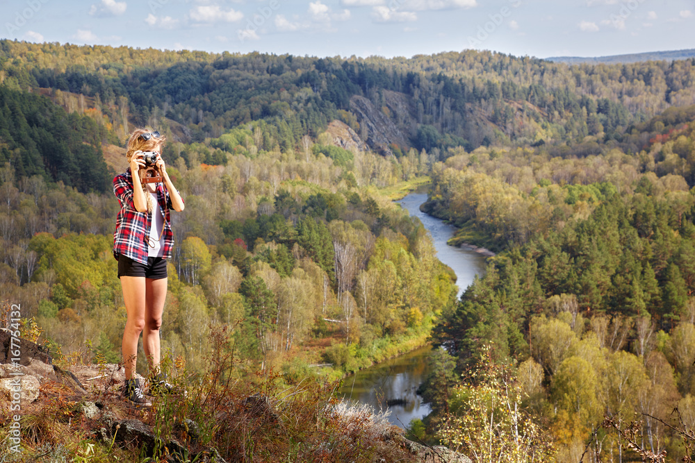 Young blonde woman tourist   on a cliff taking pictures of the autumn landscape with the river Berd on old film camera
