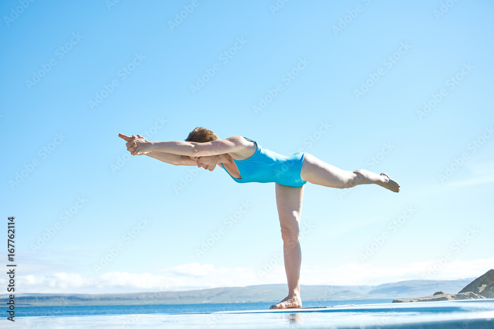 Fototapeta premium Cheerful sportive woman in yoga pose on edge of pool looking away on background of sea, Iceland.