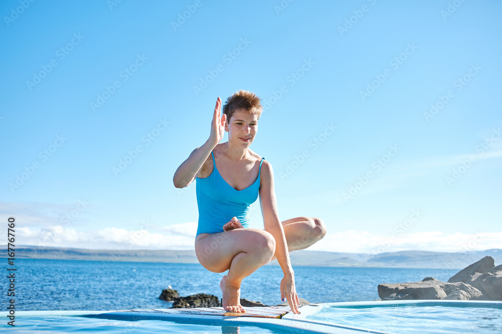 Cheerful sportive woman in yoga pose on edge of pool looking away on background of sea, Iceland.