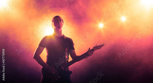 Silhouette of guitarist with a guitar on stage on orange background with smoke and spotlights 