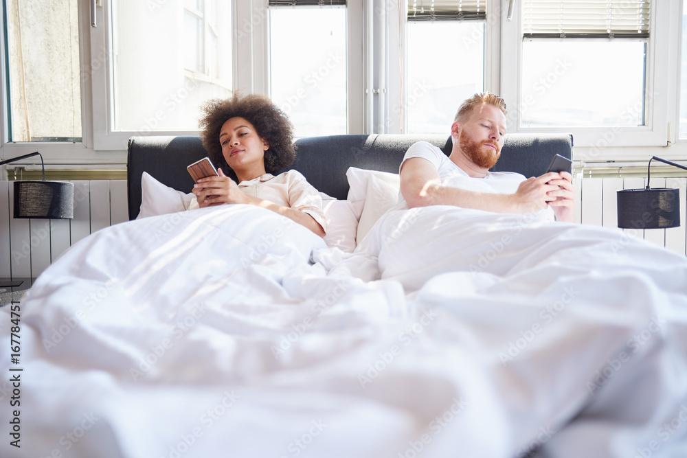 Young couple using mobile phones and lying in bed while ignoring each other