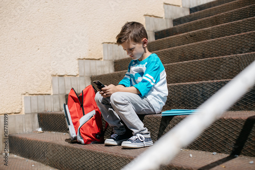 Child sitting outside school and playing games on his cell phone. Schoolboy taking a break from school and browsing his mobile.
