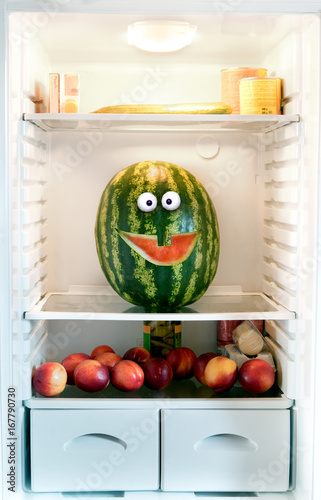 Watermelon on a shelf in the refrigerator. Cheerful funny watermelon with carved a smile looks out of fridge.