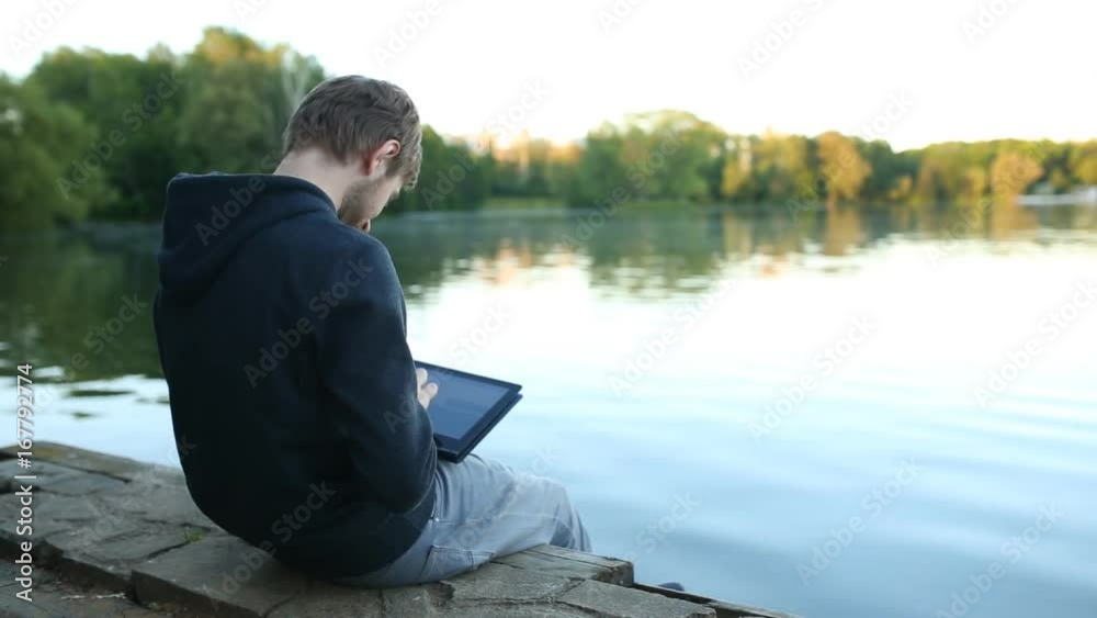 Man freelancer working on the lake with a tablet computer, image at sunset. The lights of a sun.