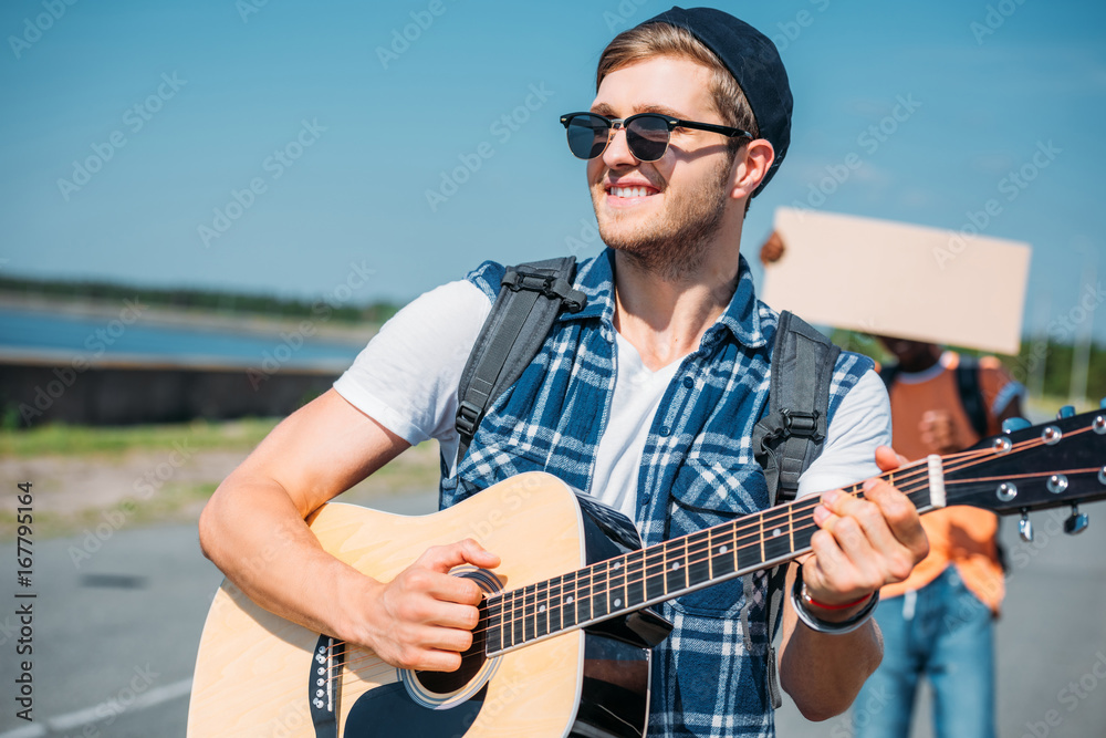selective focus of caucasian man playing guitar while african american friend with empty cardboard hitchhiking behind