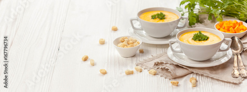 Pumpkin cream soup with croutons, raw fresh pumpkin pieces and herbs on a white rustic wooden background, Autumn concept. Long web format, banner photo