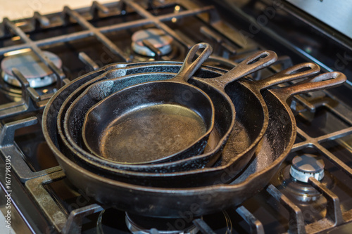 An iron skillet collection, ready to start cooking, stacked on top of a stovetop with a selective focus on the edge of the pans.