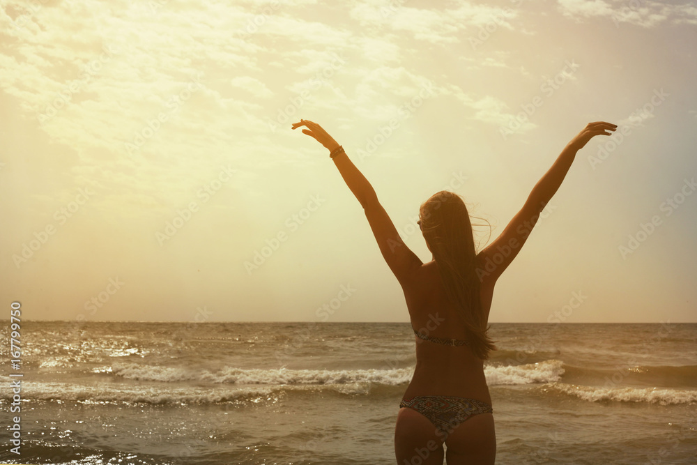 Young girl from the back raised her hands up against the sunset background next to the sea. Hands like the wings of a bird. Freedom, rest, travel, vacation. Silhouette