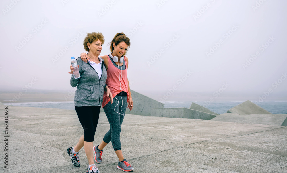 Senior woman and young woman walking outdoors by sea pier - obrazy, fototapety, plakaty 