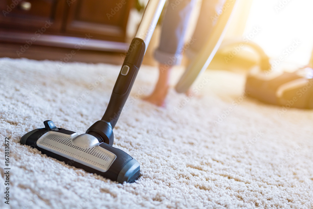 Woman using a vacuum cleaner while cleaning carpet in the house. - obrazy, fototapety, plakaty 