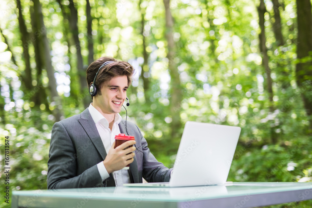 Manager with headset sitting at the office desk with laptop computer, drink a cup of coffee in call center in green forest park. Business concept. Manager with headset sitting at the office desk with laptop computer, drink a cup of coffee in call center in green forest park. Business concept.
