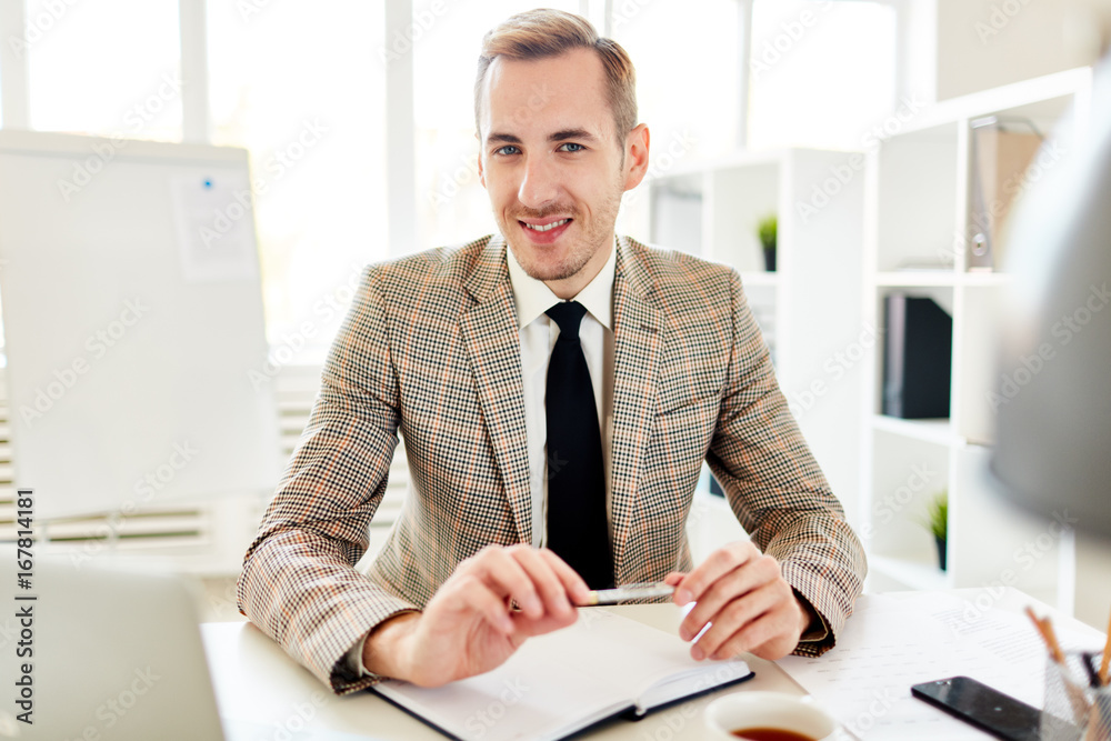 Waist-up portrait of handsome young entrepreneur looking at camera with toothy smile while sitting at desk, interior of modern office with panoramic windows on background Waist-up portrait of handsome young entrepreneur looking at camera with toothy smile while sitting at desk, interior of modern office with panoramic windows on background