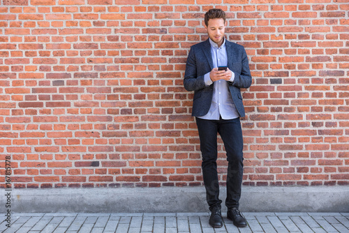 Businessman sms texting phone app in city street on brick wall background. Business man holding smartphone in smart casual wear standing. Urban young professional lifestyle. © Maridav