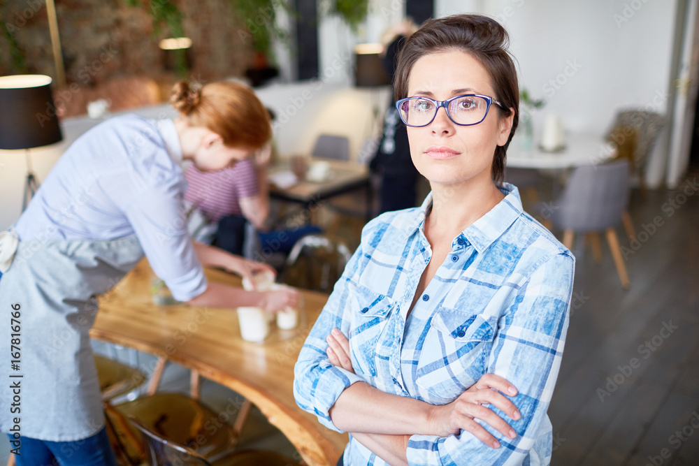 Obraz premium Portrait of confident restaurant owner posing for photography with arms crossed, busy red-haired waitress wrapped up in work seen on background