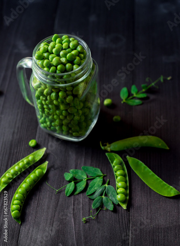 Bunch of fresh mature pods of green peas tied with a rope near full glass jar with green peas on black wooden background. Bio healthy food. Green peas, pods, pea leaves and jar on wooden table