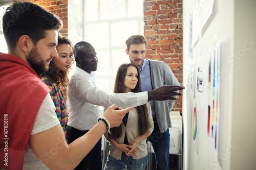 Collaboration is a key to best results. Group of young modern people in smart casual wear planning business strategy while young man pointing at infographic displayed on white wall in the office photo