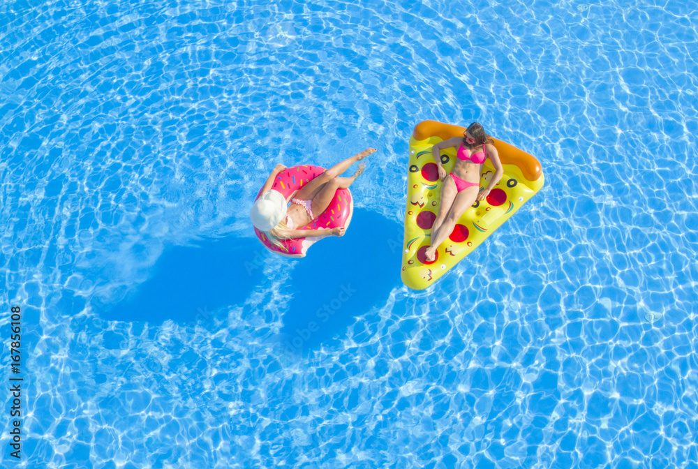 AERIAL TOP DOWN Relaxed girls in pink bikini swimsuits lying on fun inflatable pizza and flamingo floating pillows on water. Girlfriends on summer vacation enjoying colorful floats in swimming pool